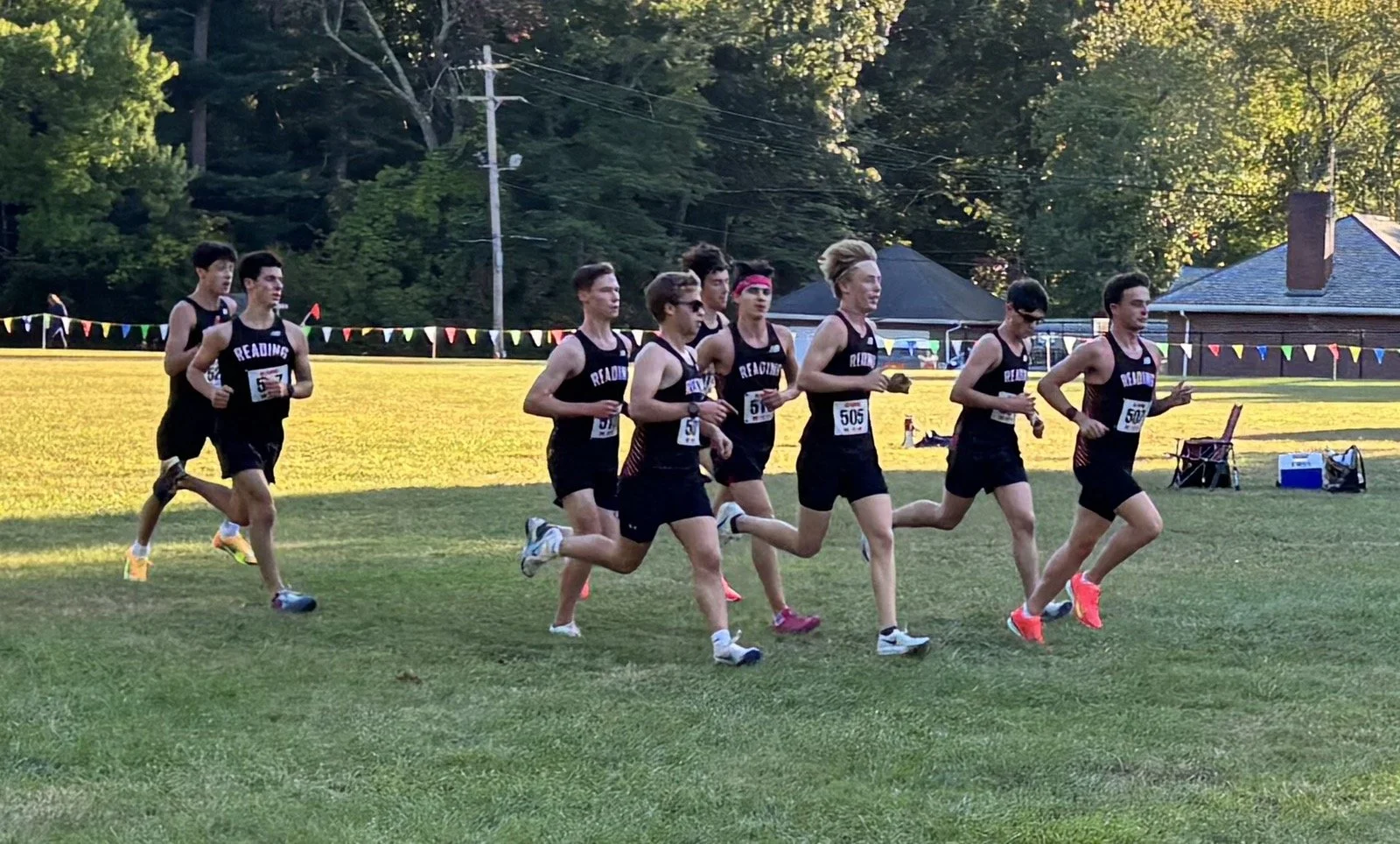 Image 1 (boys' team): Reading boys' cross-country runners competing in a pack during a Middlesex League meet, wearing black uniforms on a grass course lined with spectators and pennant flags.