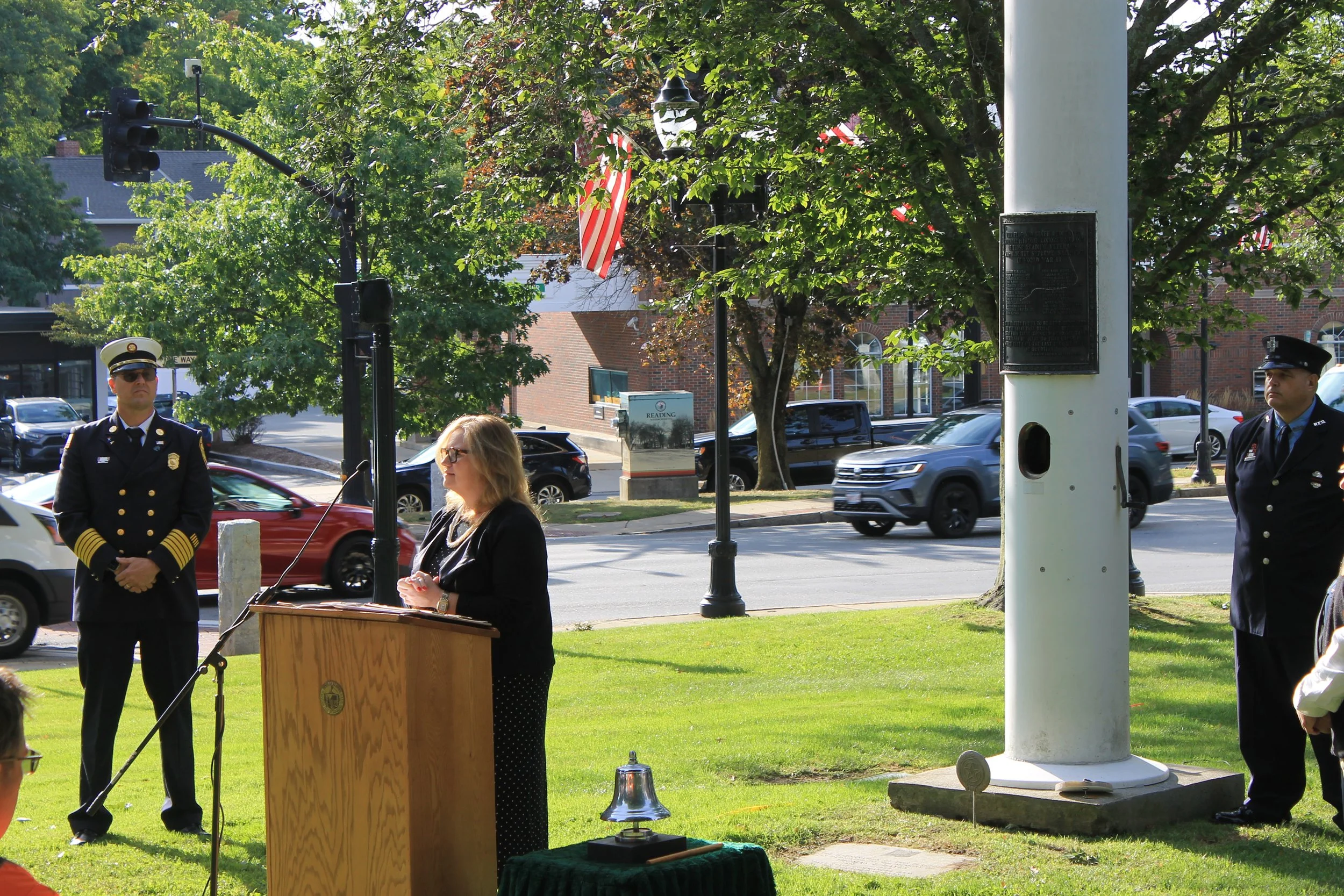 A woman in dark clothing speaks at a podium on the town common, with fire officials and a ceremonial bell nearby.