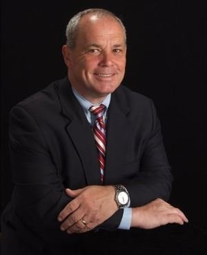Portrait of Terry Kennedy, a smiling middle-aged white man with short gray hair, wearing a dark suit and red striped tie, arms crossed, against a black background.