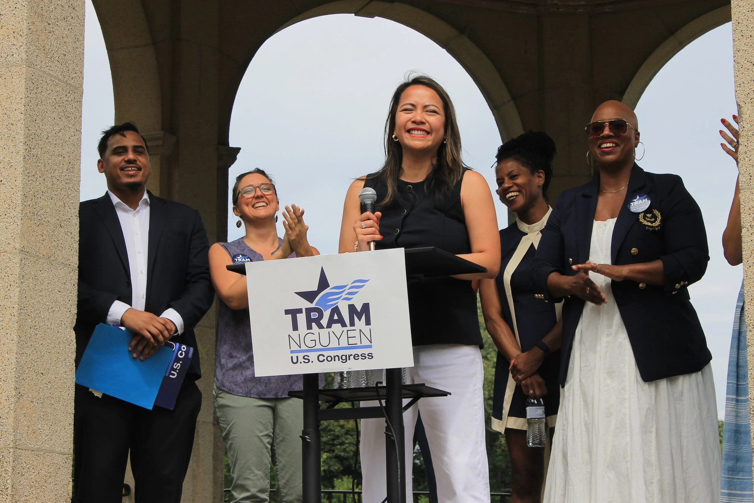 Tram Nguyen smiles while speaking at the podium, flanked by supporters clapping under the Salem Common gazebo.
