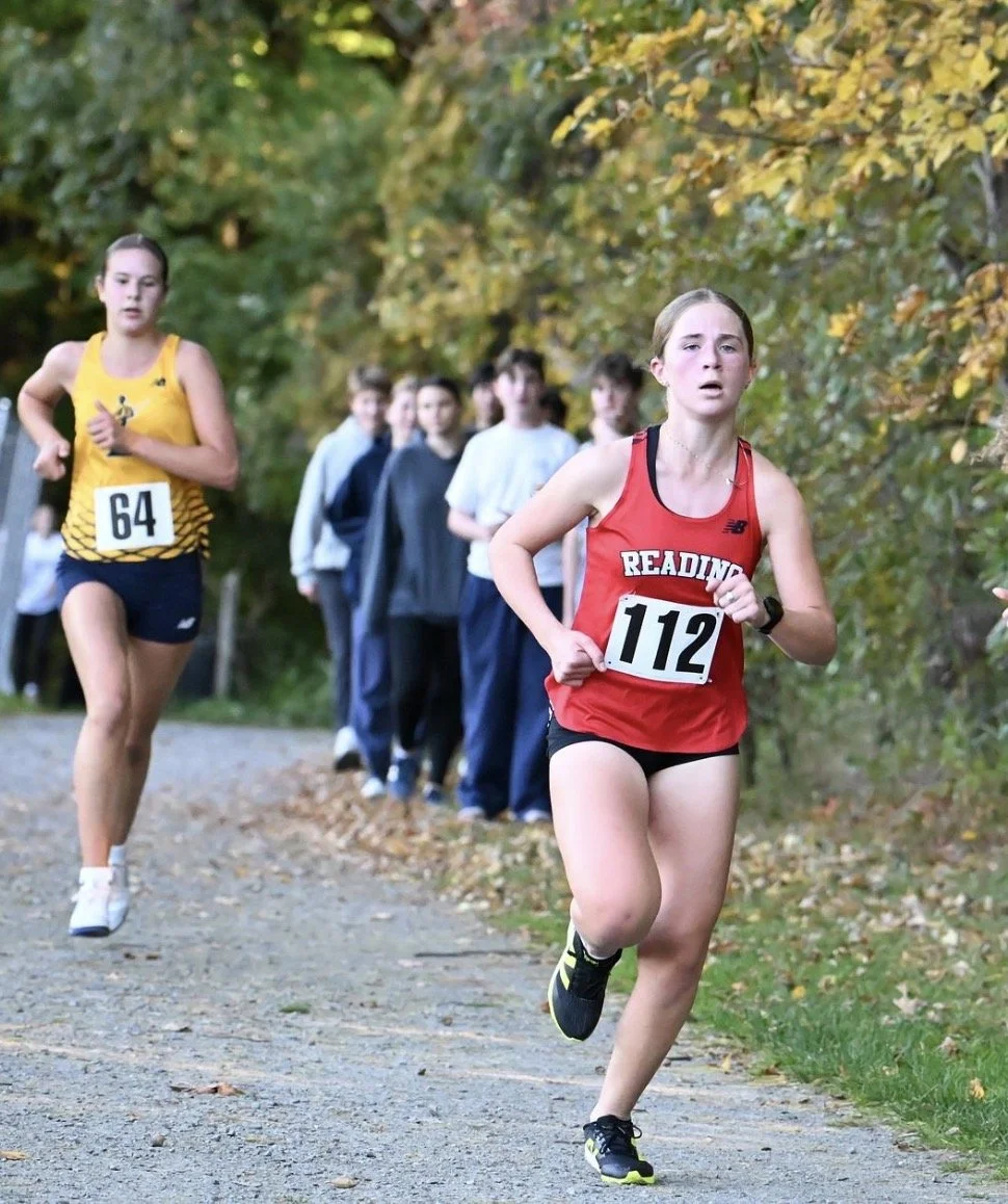 Reading girl in a red uniform and bib 112 runs on a gravel path ahead of a runner in yellow, with spectators and fall leaves behind them.