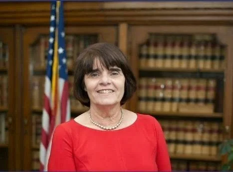 Portrait of Marian Ryan, a smiling woman with short dark hair, wearing a red dress and pearl necklace, in front of an American flag and shelves of law books.