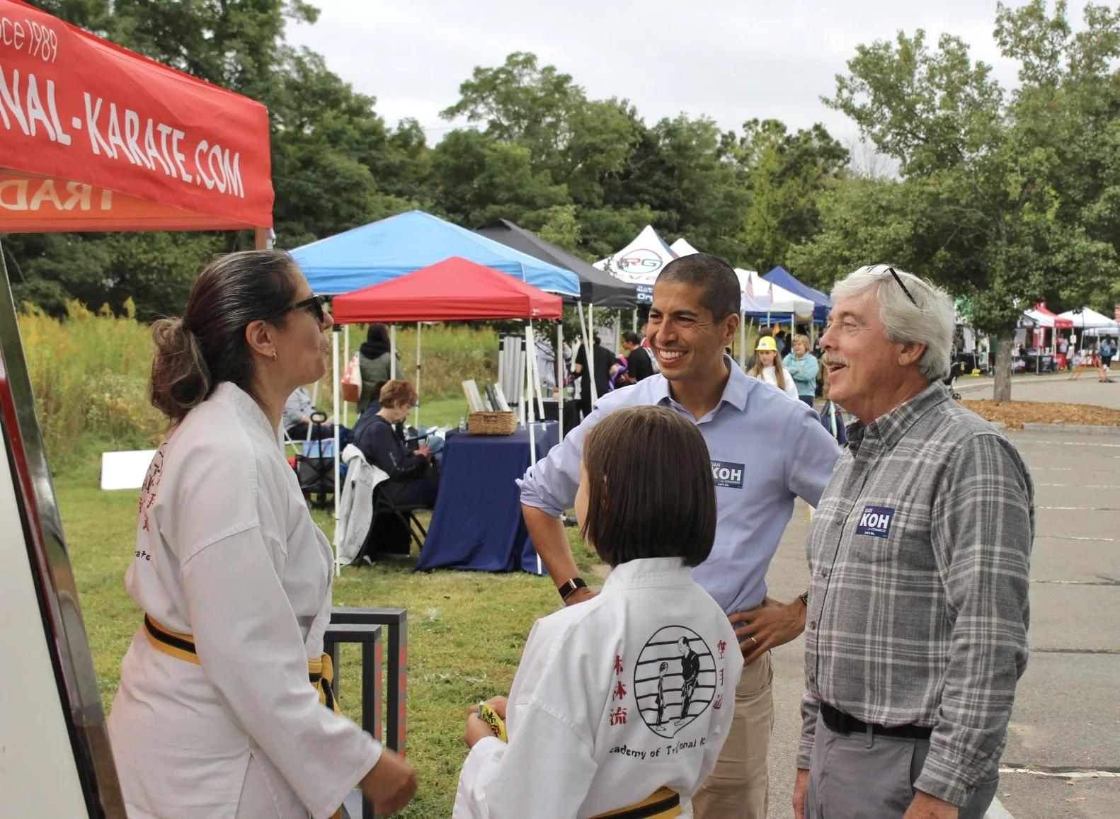 Dan Koh, wearing a Koh campaign sticker, laughs with an older man and talks with two karate students and an instructor at an outdoor community event.