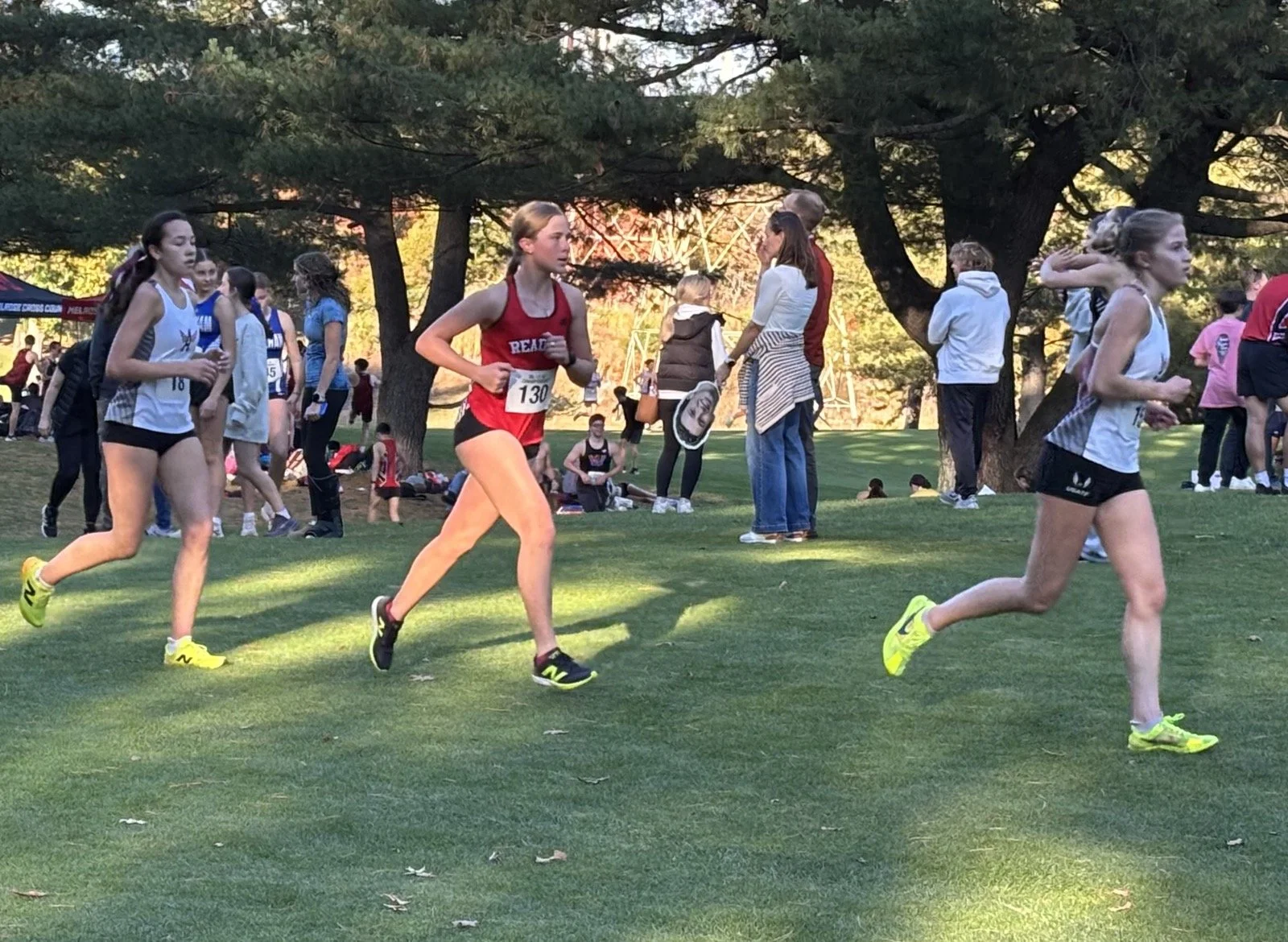 A Reading girls cross-country runner in a red uniform and bib 130 runs between two runners in white uniforms, with spectators and fall trees in the background.