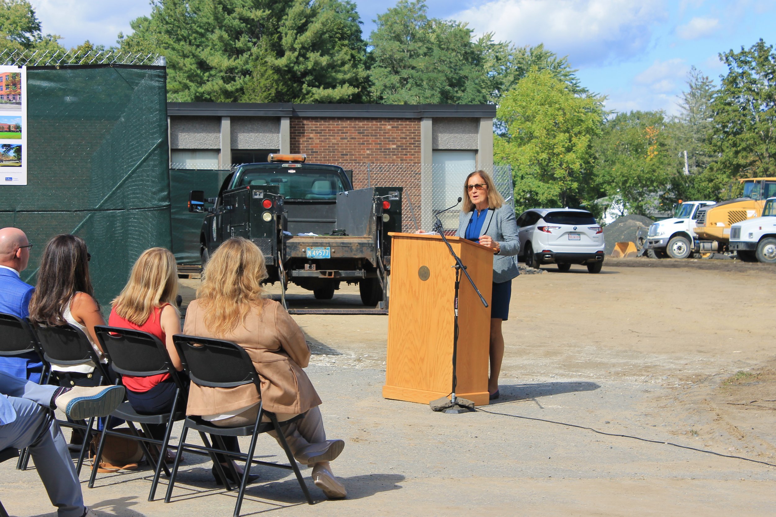 A speaker in a gray blazer speaks at an outdoor podium to a seated crowd, with a brick building and truck in the background.