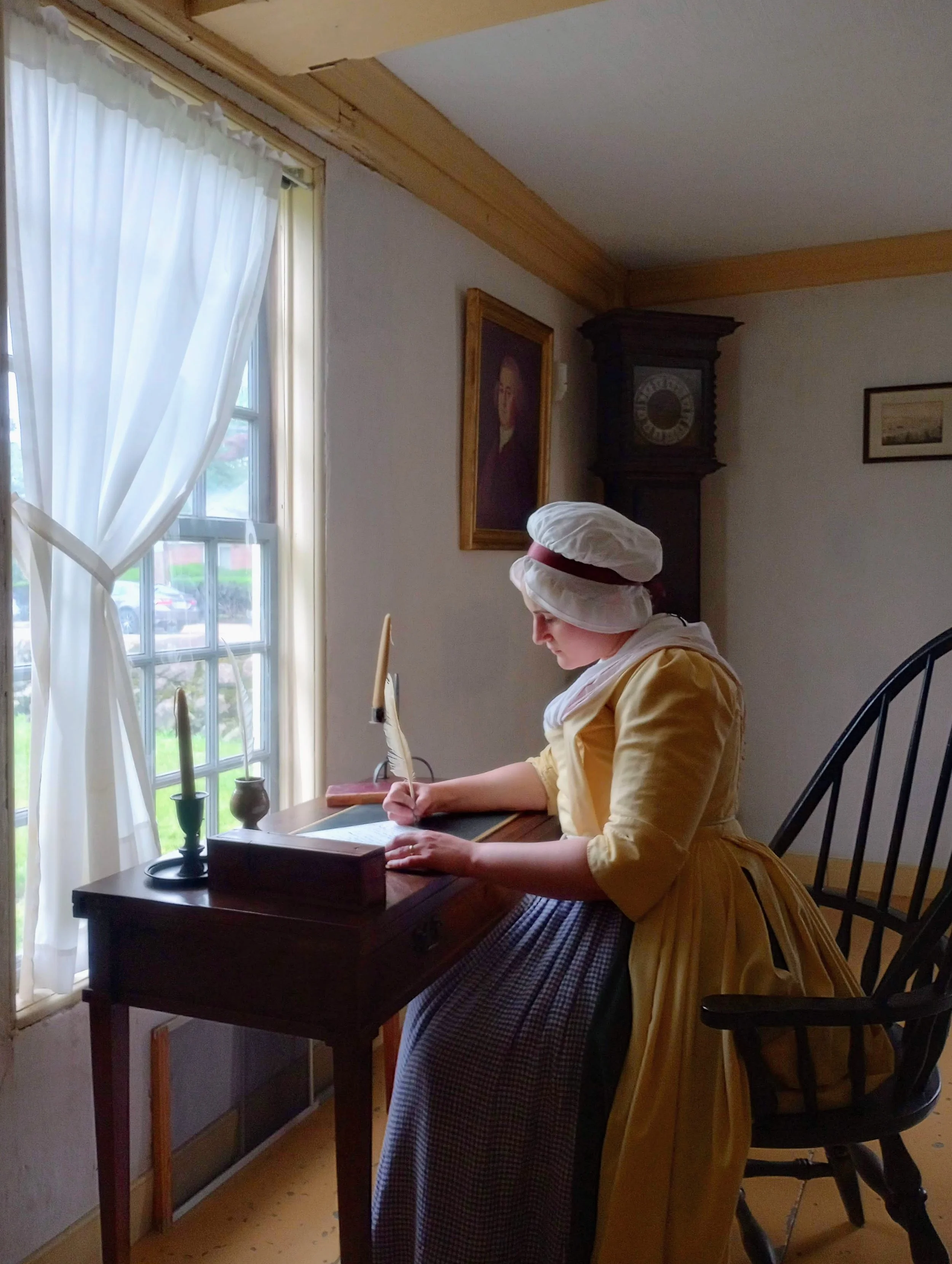 A woman in colonial-era dress and cap writes with a quill pen at a period writing desk in a historic room.