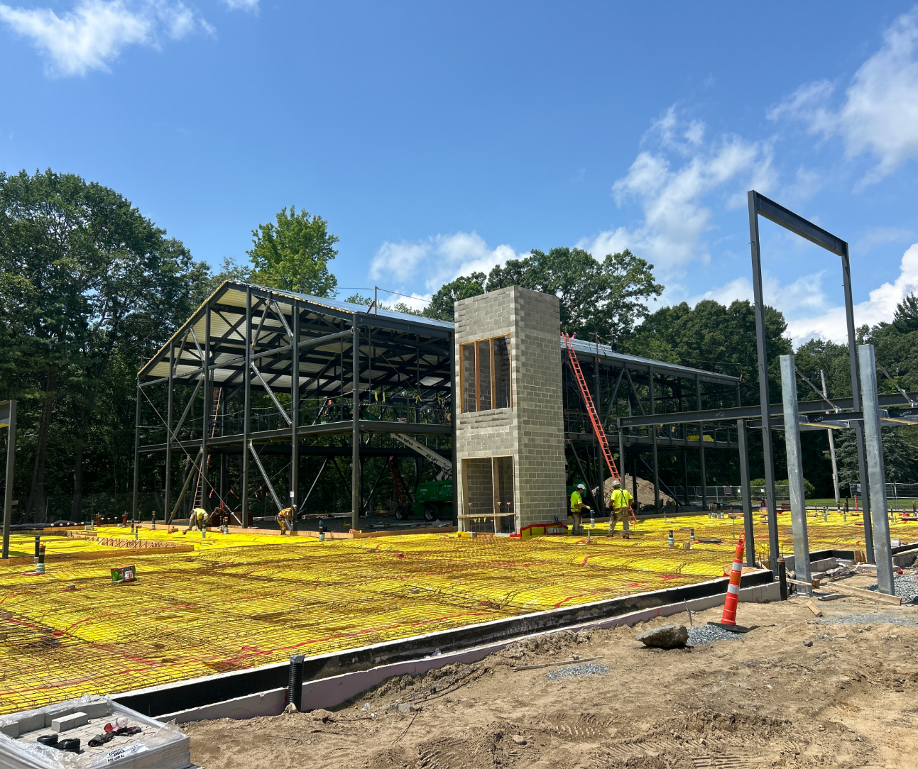 A steel building frame under construction on a sunny day, with a concrete block elevator shaft at the center and yellow reinforcing mesh covering the ground floor in the foreground.