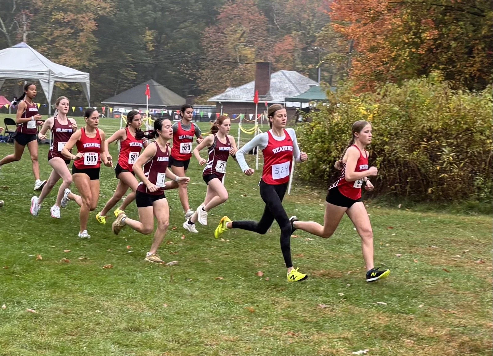 Reading girls in red and Arlington girls in maroon run across a grass field past a pavilion, with orange and yellow fall foliage behind them. (141)