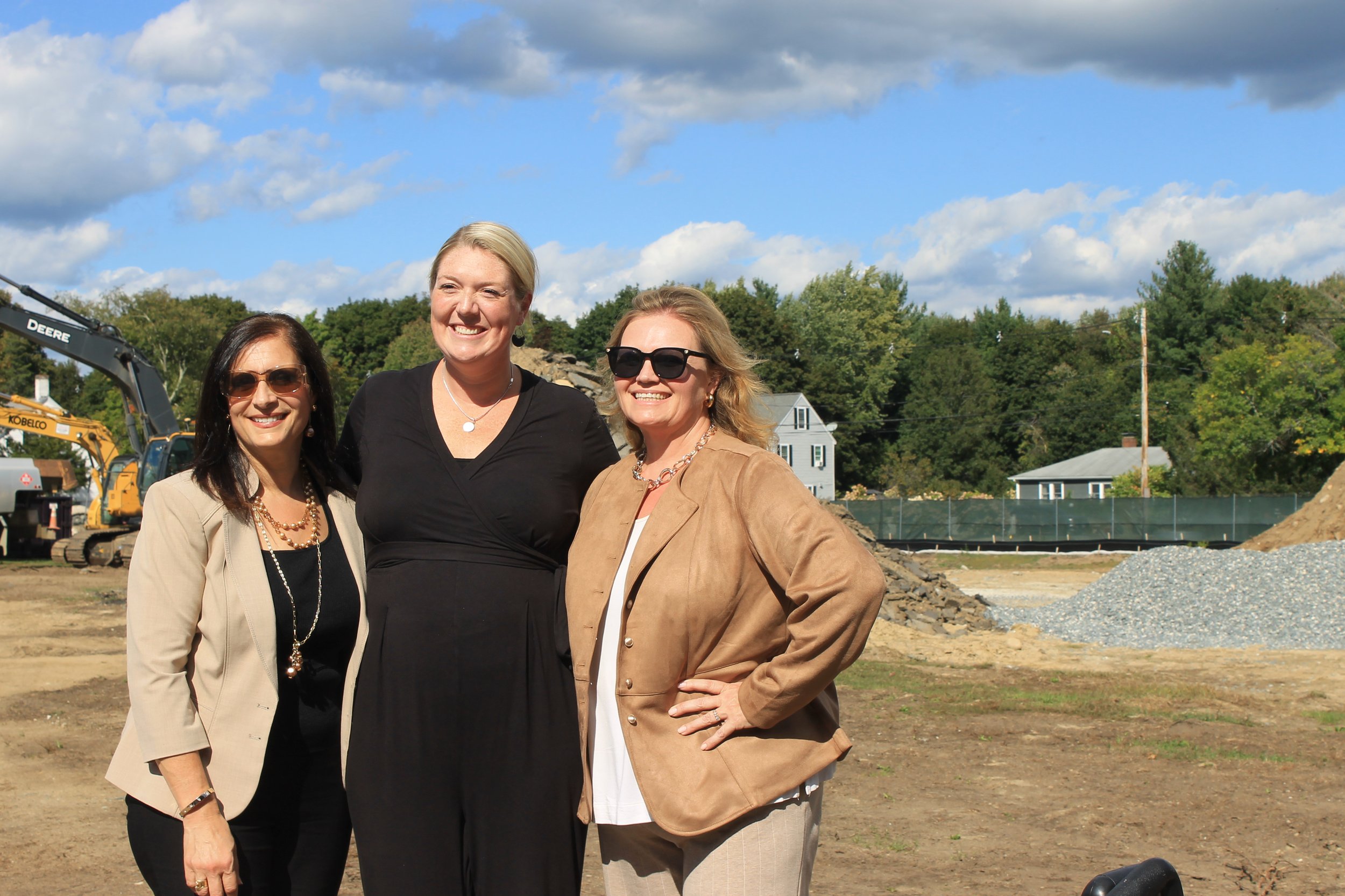 Three women stand together and smile for a photo at the Killam School construction site.
