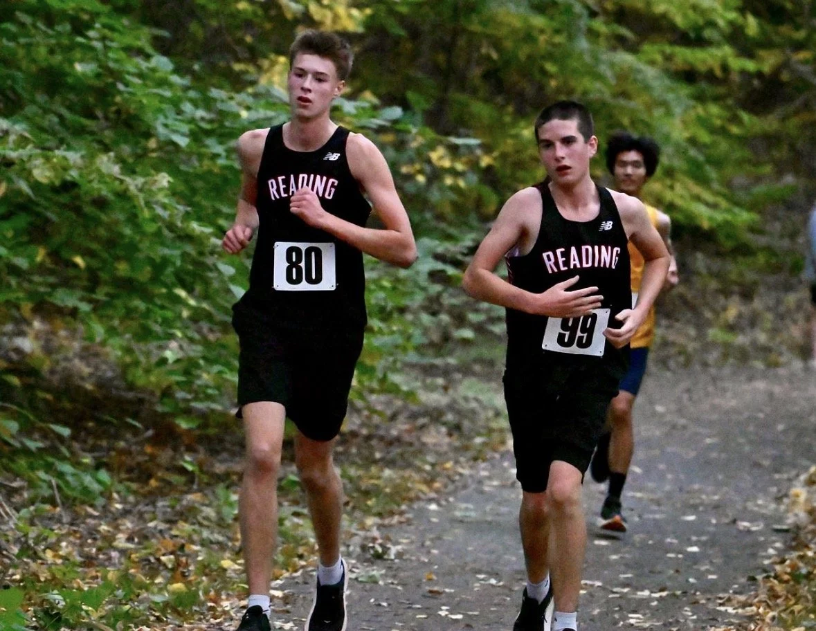 Two Reading boys in black uniforms, bibs 80 and 99, run along a wooded path as a runner in yellow follows.