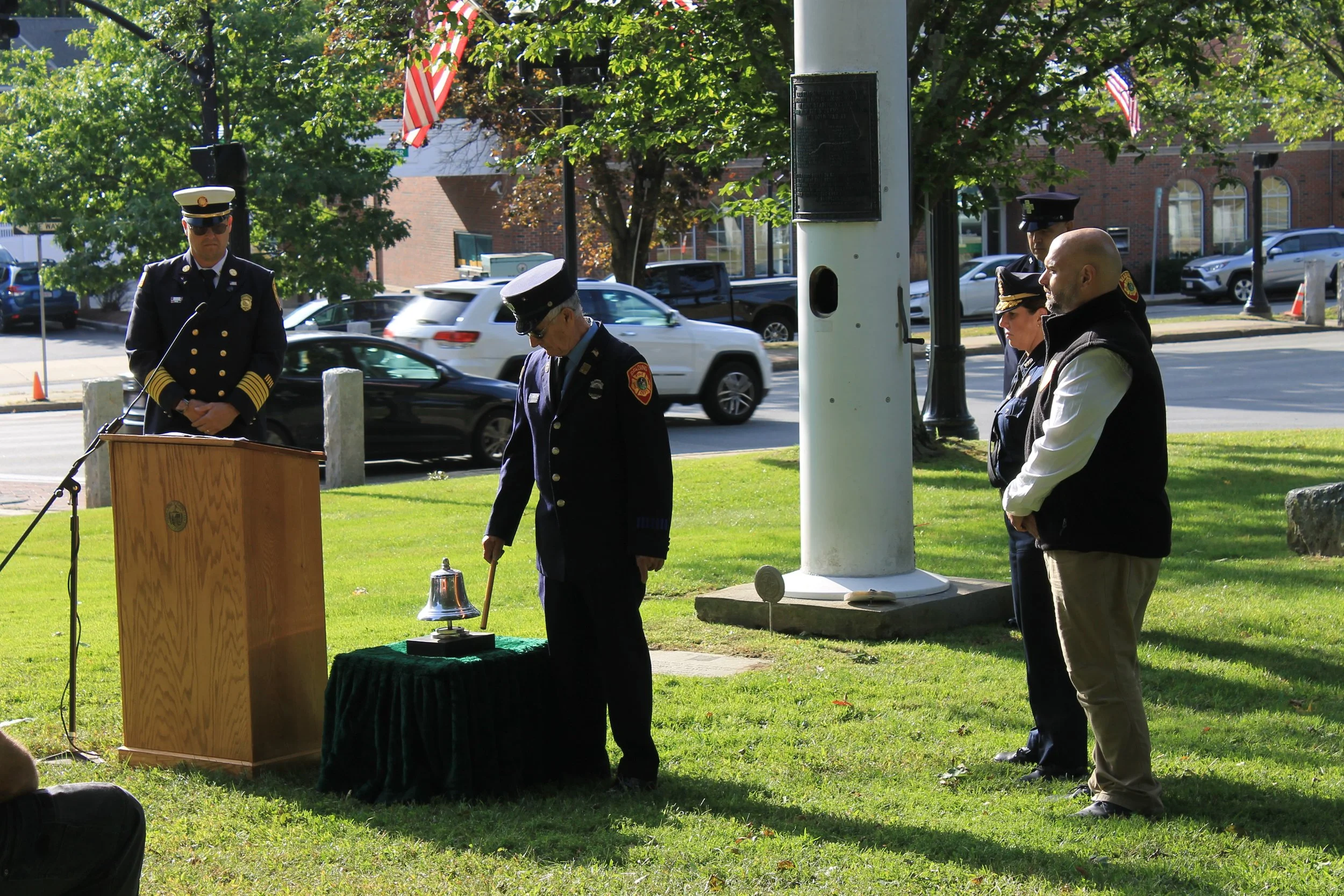 Reading fire chief stands at a podium as a firefighter in dress uniform tolls a ceremonial bell on a draped table.