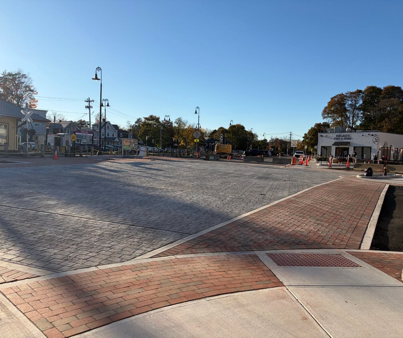 A wide gray paved intersection with brick sidewalk corners, orange cones, and concrete barriers, with a railroad crossing signal and a white restaurant building in the distance.