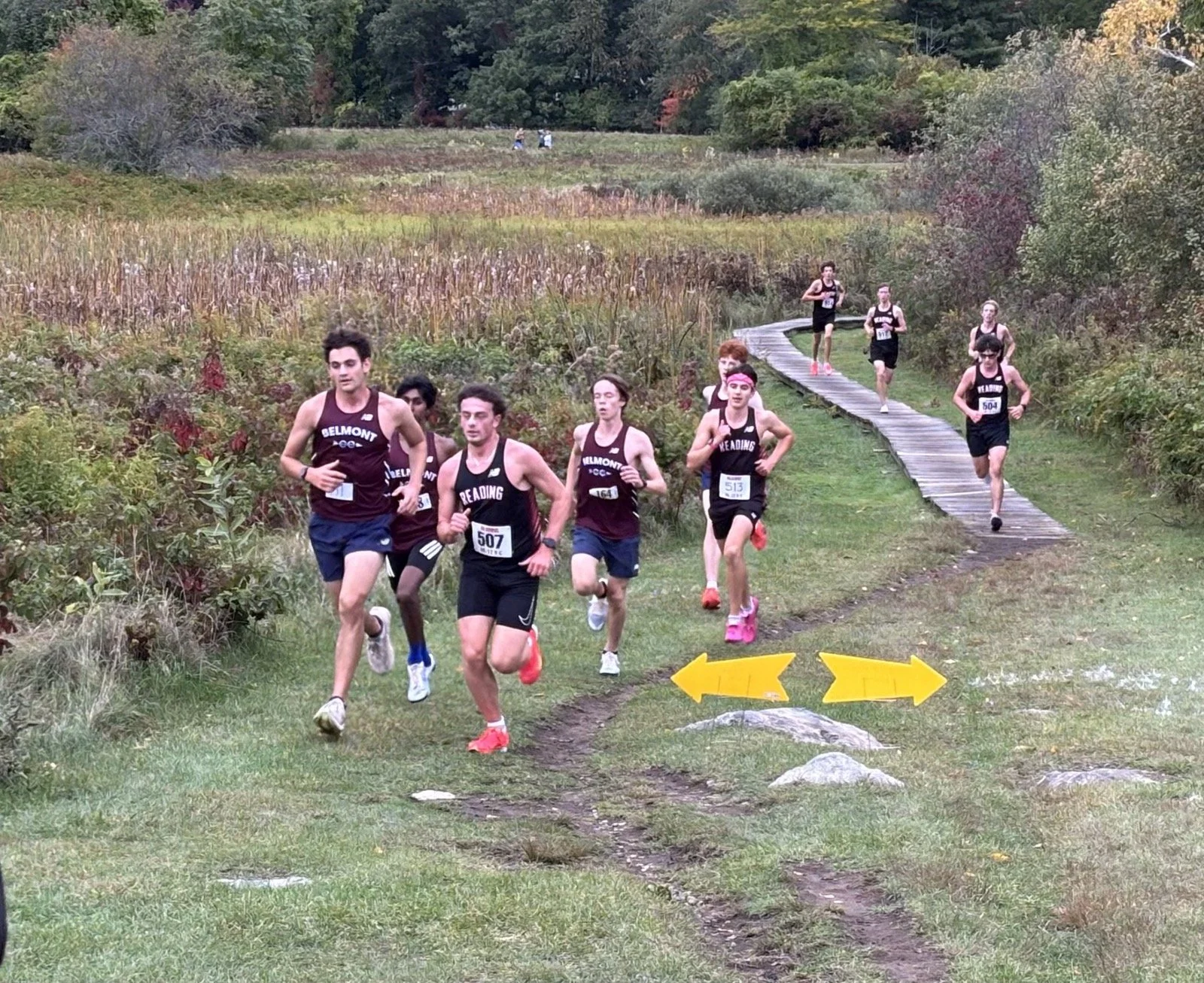 Reading boys' cross-country runners in black jerseys race alongside Belmont runners in maroon on a grassy trail beside a wooden boardwalk and marsh grass during a Middlesex League meet.