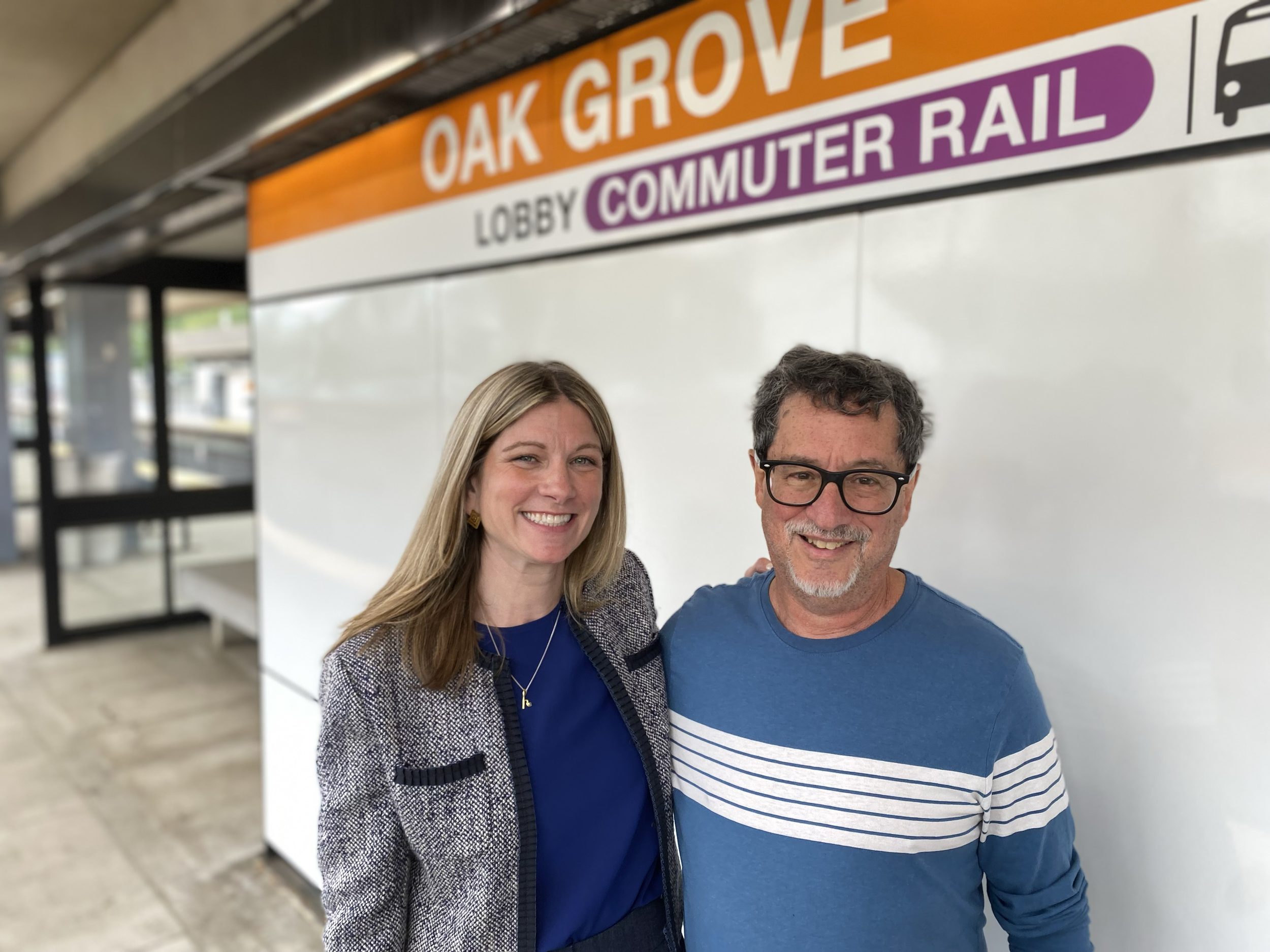 Kate Lipper-Garabedian stands with a man in a blue striped shirt beneath an Oak Grove Commuter Rail station sign.