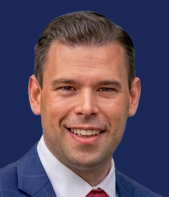 Campaign headshot of Joe Markey, a man with short dark hair smiling against a navy backdrop, wearing a blue suit, white shirt, and red tie.
