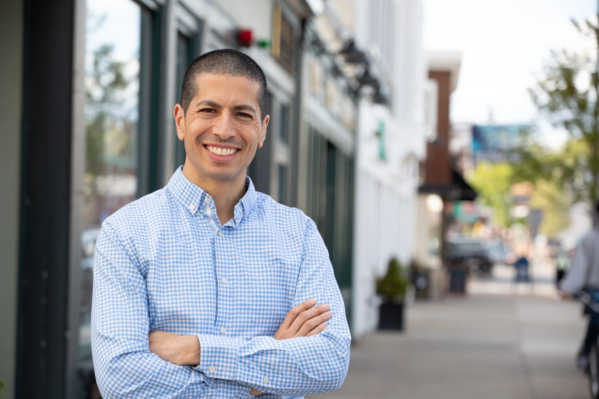 Dan Koh, wearing a blue checkered shirt, smiles with arms crossed on a sidewalk in front of storefronts.
