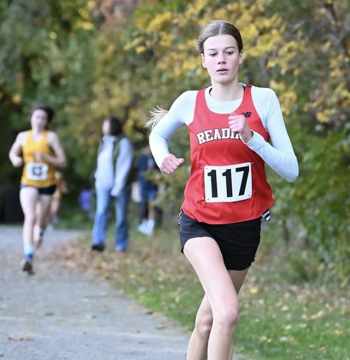 Reading girl in a red uniform and bib 117 runs on a gravel path lined with fall foliage as a runner in yellow follows behind.