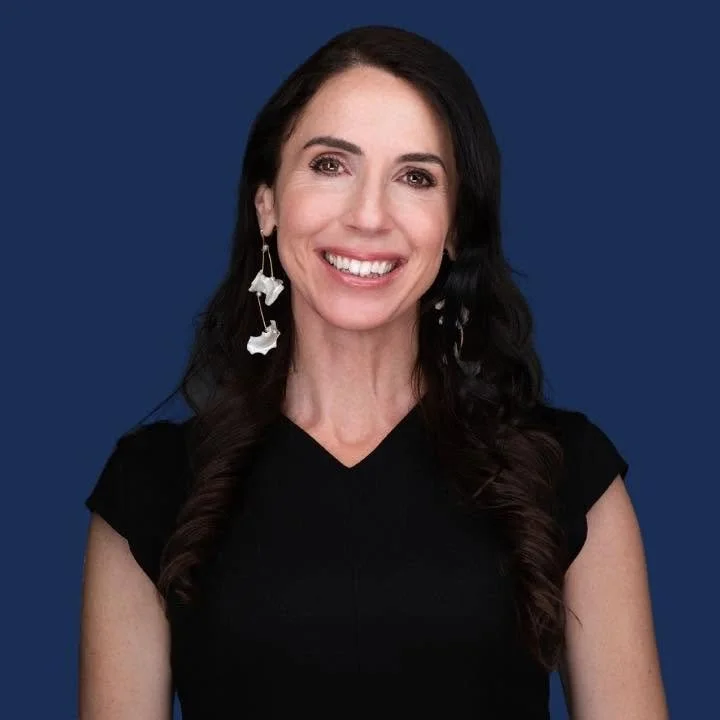 Portrait of Diana DiZoglio, a smiling woman with long dark curly hair and drop earrings, wearing a black top, against a navy background.