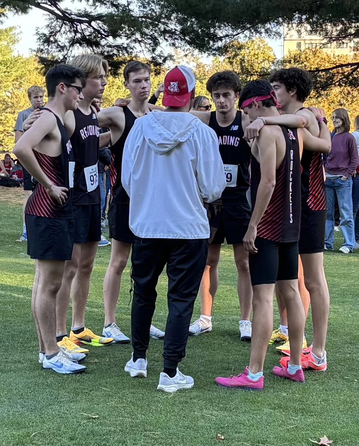 Reading boys cross-country runners huddle with arms around each other as a coach in a white jacket and red cap, back to the camera, speaks to them in a park.