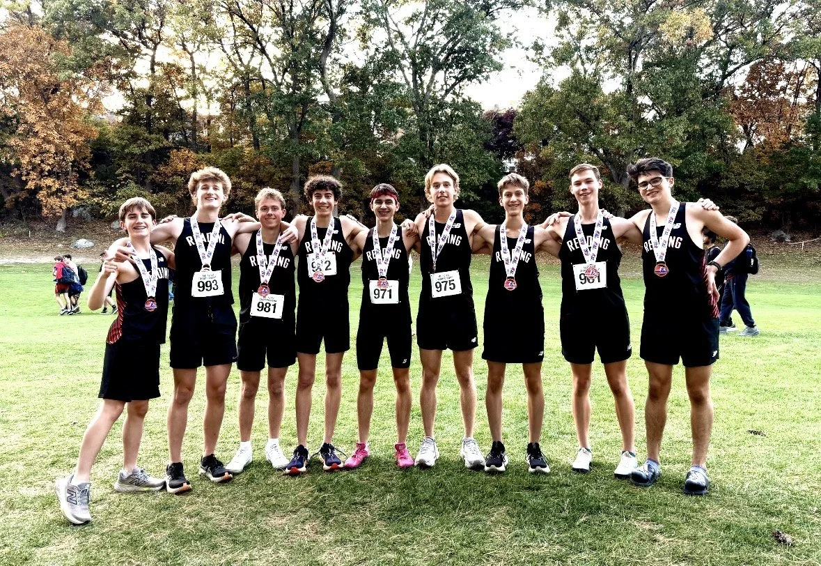 Nine Reading boys cross-country runners in black uniforms and medals stand side by side with arms around each other on a grassy field, with trees in fall color behind them.