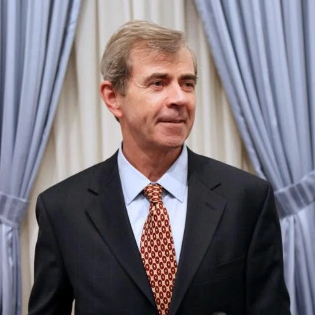 Portrait of Bill Galvin, a middle-aged white man with gray hair, wearing a dark suit, light blue shirt, and patterned red tie, standing in front of pale curtains.