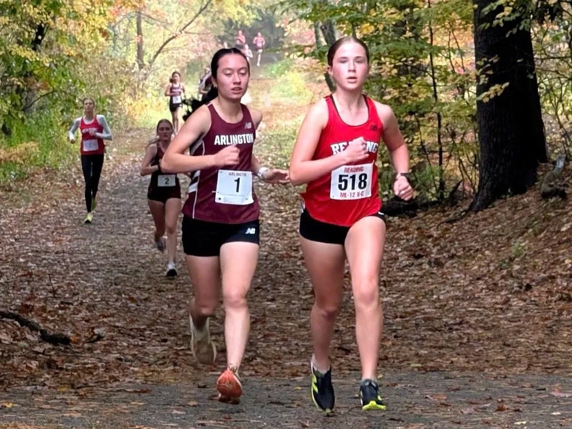 An Arlington runner in maroon and a Reading runner in red, bib 518, run side by side on a leaf-covered trail, with more runners behind them.
