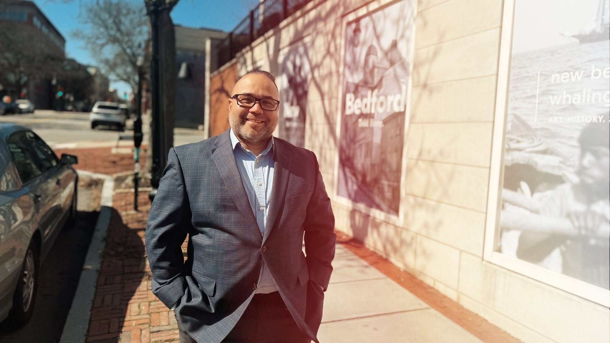 Portrait of Shawn Oliver, a smiling middle-aged man with glasses and a short beard, wearing a gray plaid blazer over a light blue shirt, standing on a New Bedford sidewalk.