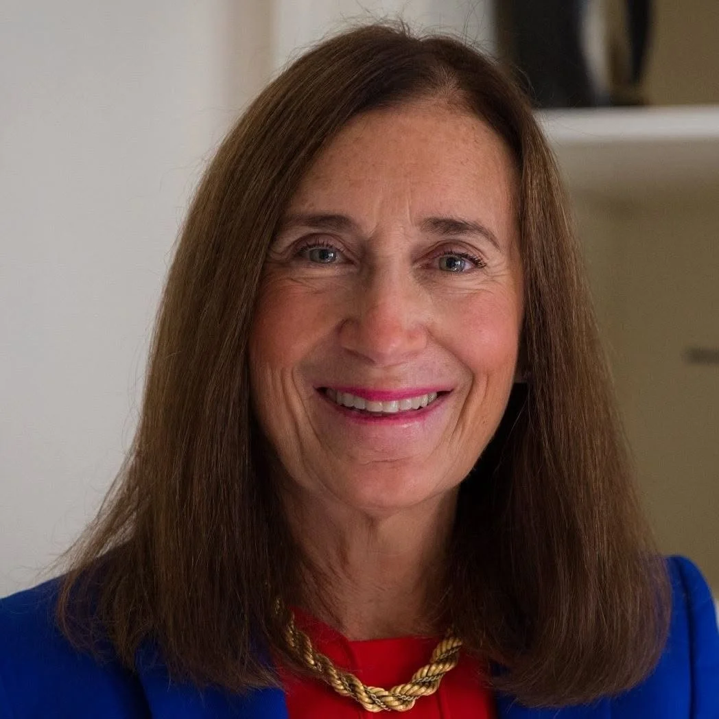 Portrait of Deb Goldberg, a smiling middle-aged white woman with long brown hair, wearing a blue blazer over a red top with a gold rope necklace.