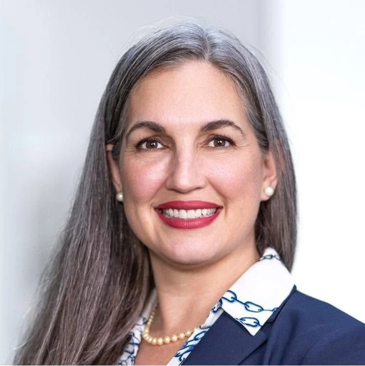 Portrait of Jaime Belsito, a smiling woman with long gray hair and red lipstick, wearing a navy blazer over a patterned collared shirt with a pearl necklace.