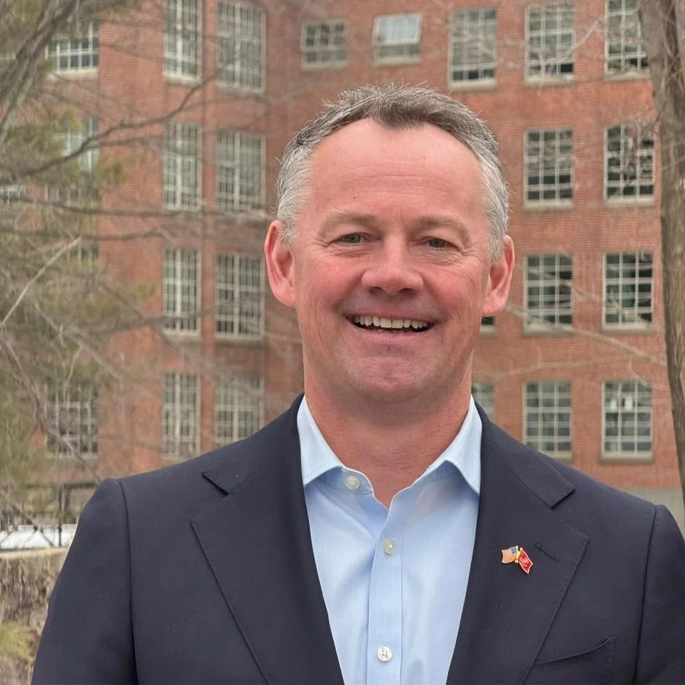 Portrait of Brian Shortsleeve, a middle-aged white man with gray hair in a navy suit and light blue open-collar shirt, in front of a brick building.