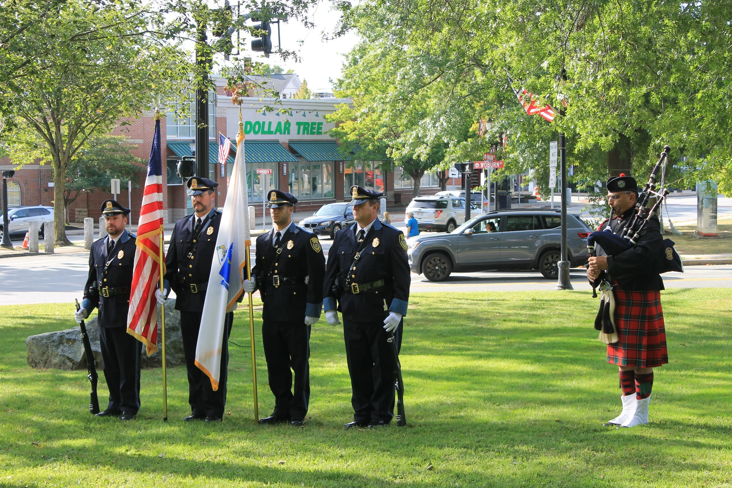 Four uniformed officers hold American and Massachusetts flags beside a bagpiper in a kilt on the town common.