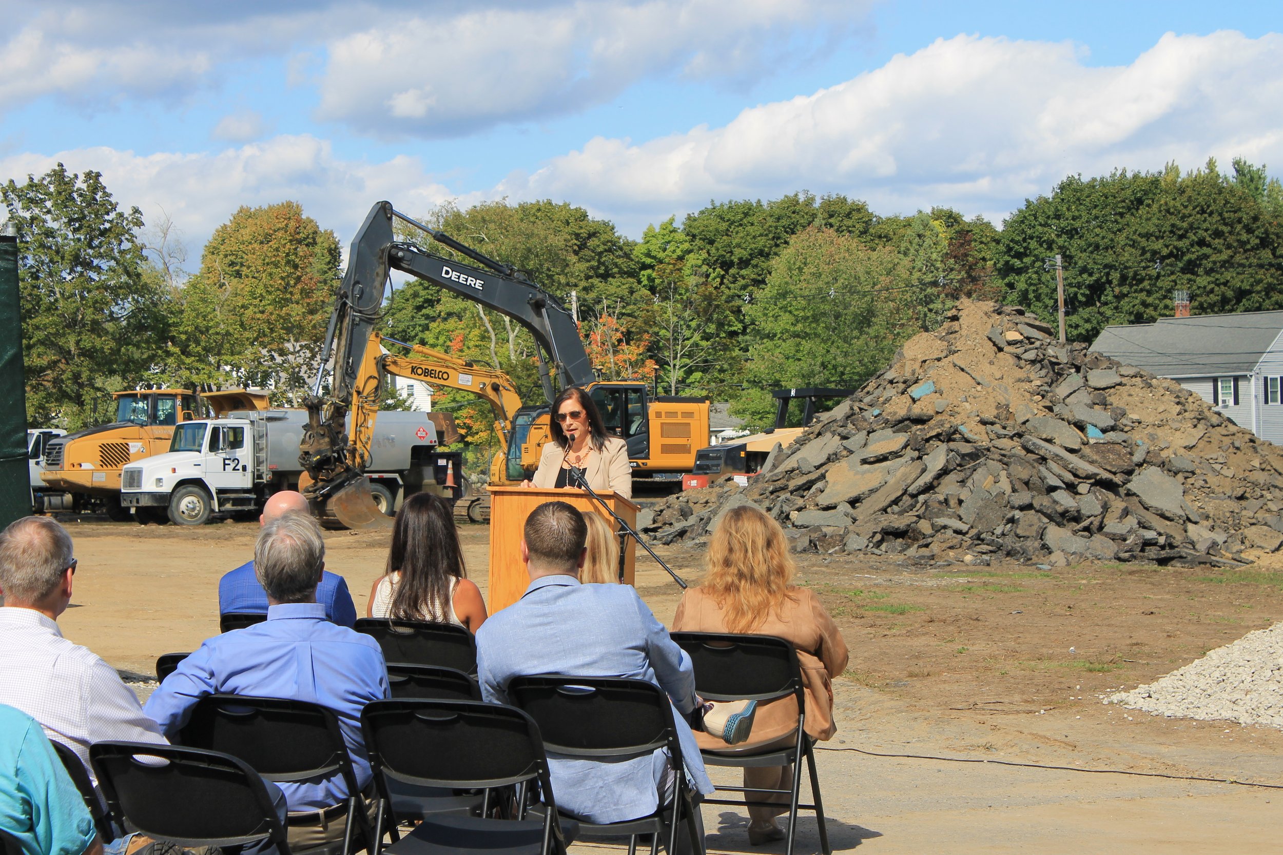 A speaker in a tan jacket speaks at an outdoor podium to a seated crowd, with an excavator and a large debris pile in the background.
