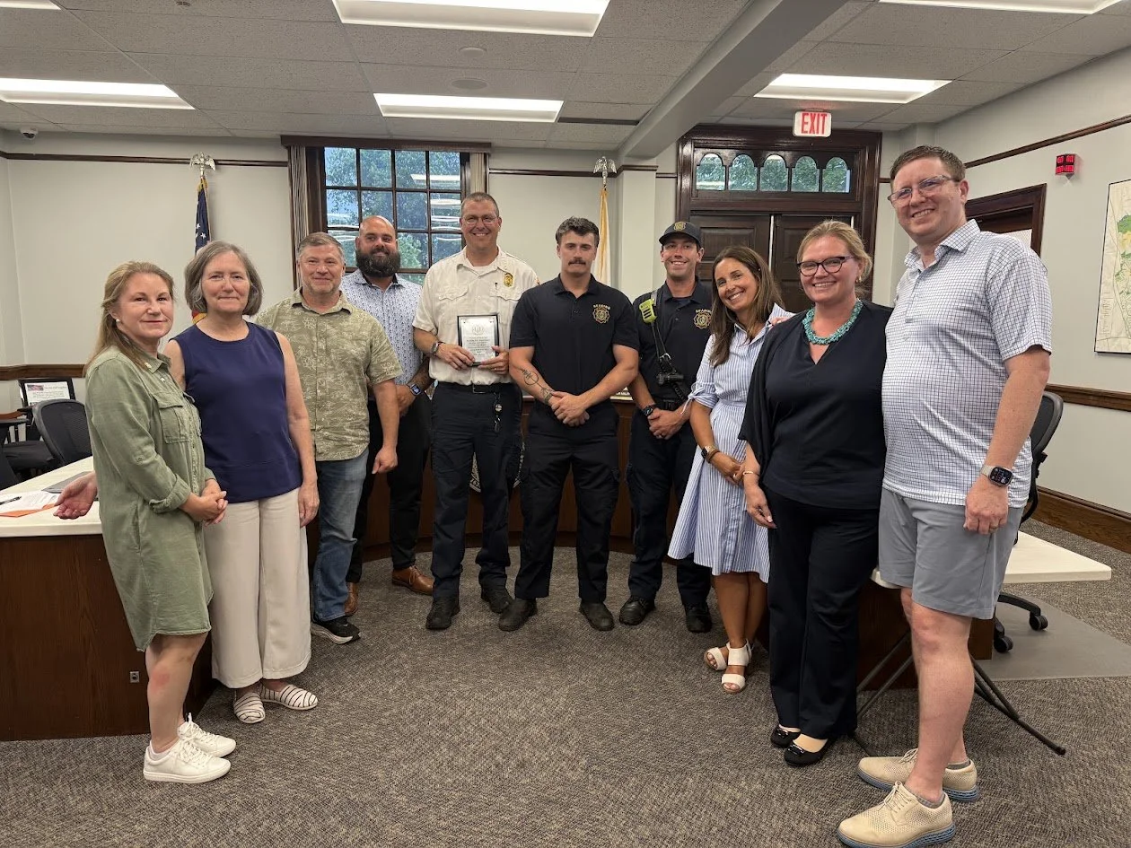 A group of about ten people pose together in the Select Board meeting room, with a fire department officer in uniform holding a plaque at the center.