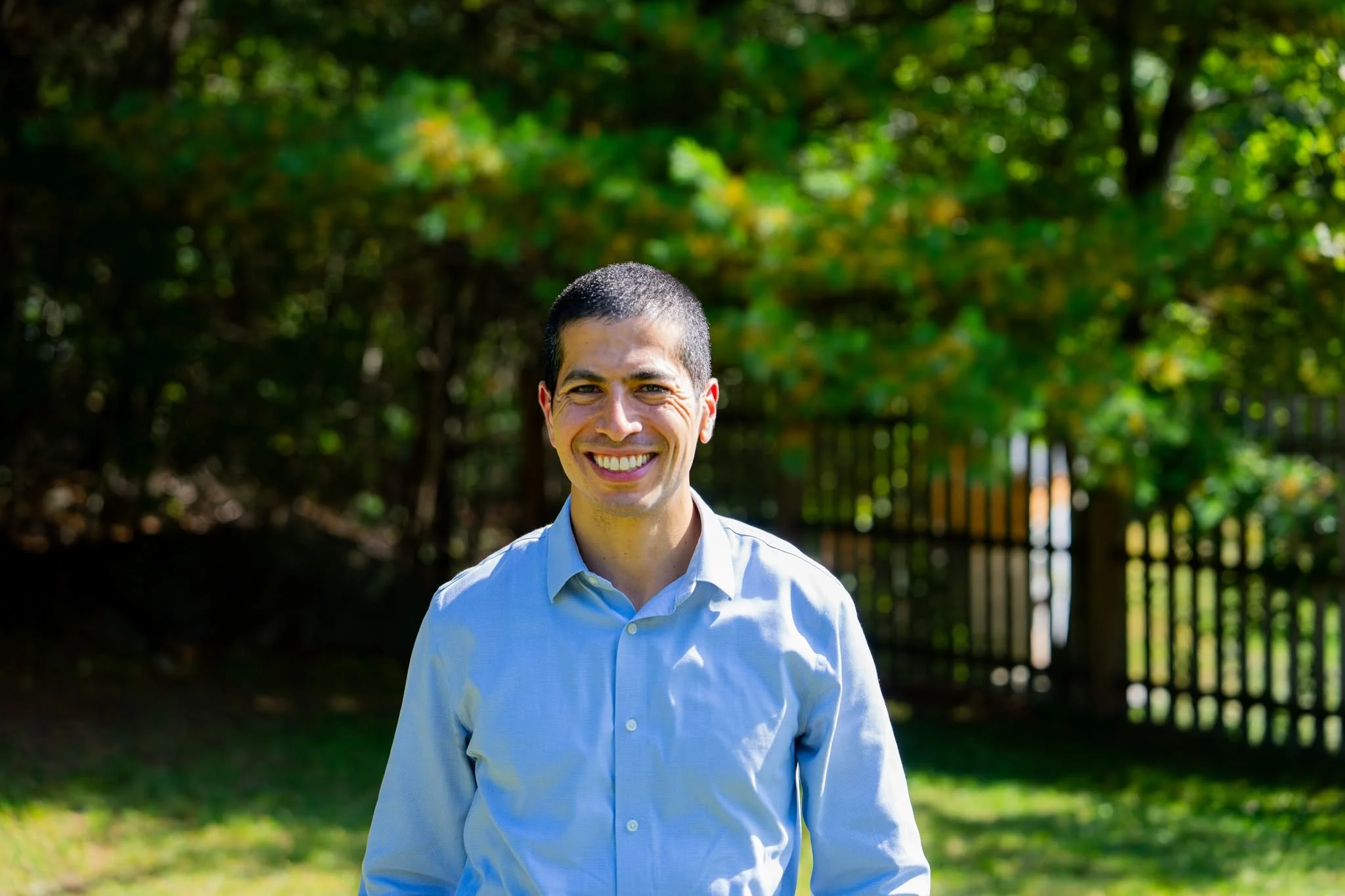 Portrait of Dan Koh, a smiling man with short dark hair, wearing a light blue button-down shirt, standing outdoors in front of green trees and a black fence.