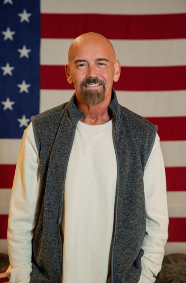Portrait of John Deaton, a bald middle-aged white man with a goatee, wearing a gray fleece vest over a cream long-sleeve shirt, standing in front of an American flag.