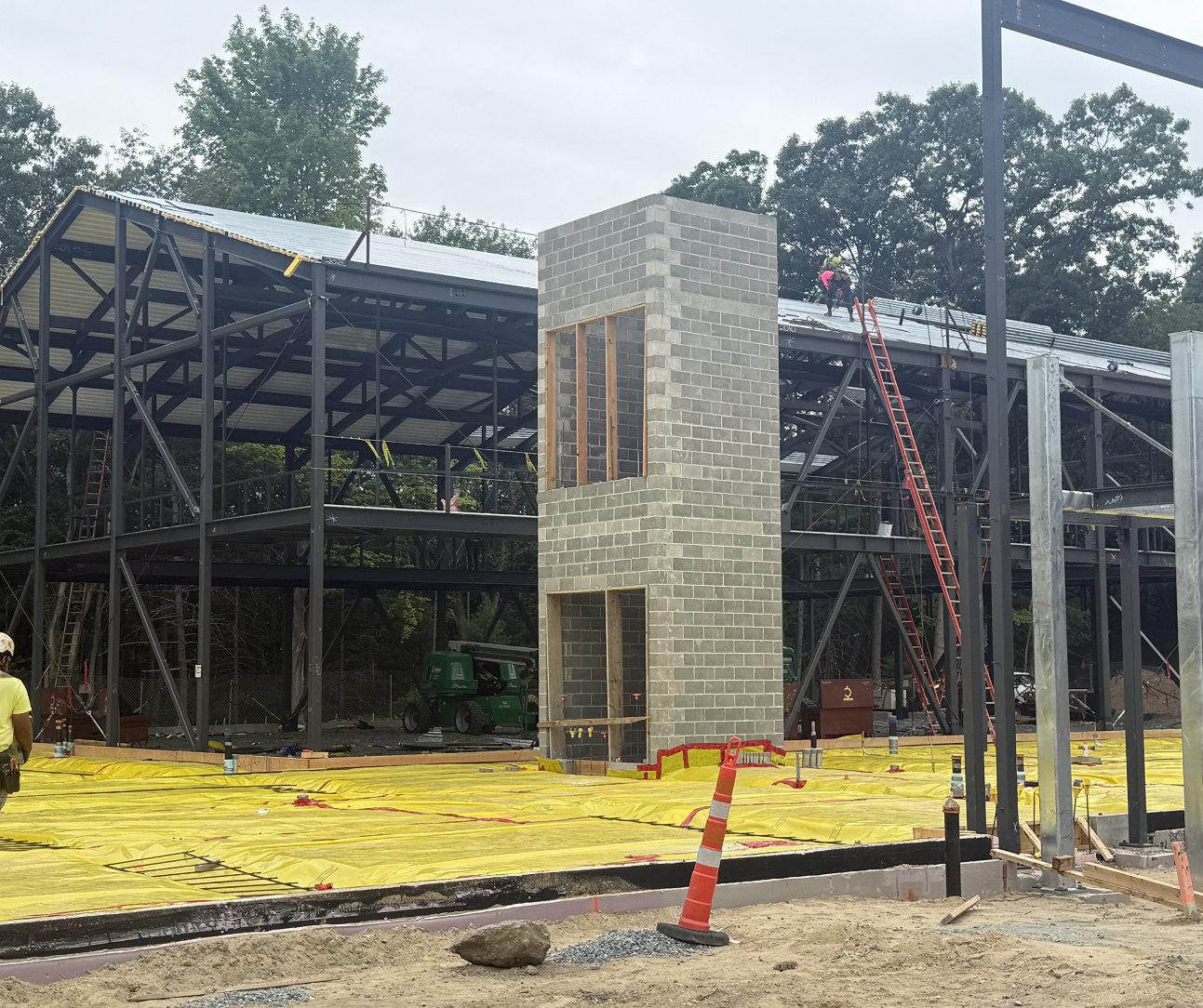 A concrete block elevator shaft stands at the center of a steel building frame, with a worker on the roof, ladders against the structure, and yellow vapor barrier sheeting covering the ground floor.