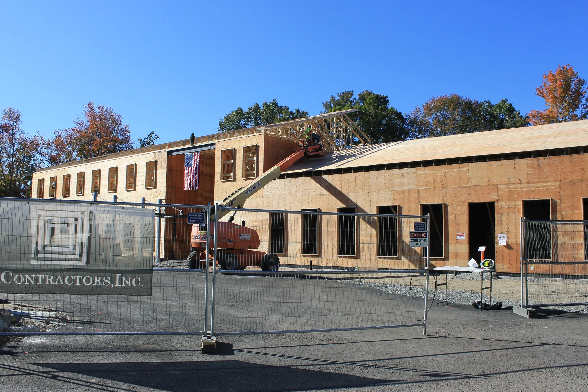 Wood-framed ReCAL building with a U.S. flag hanging from the final beam and a small evergreen on top, as a lift raises a worker at the roofline behind a construction fence.