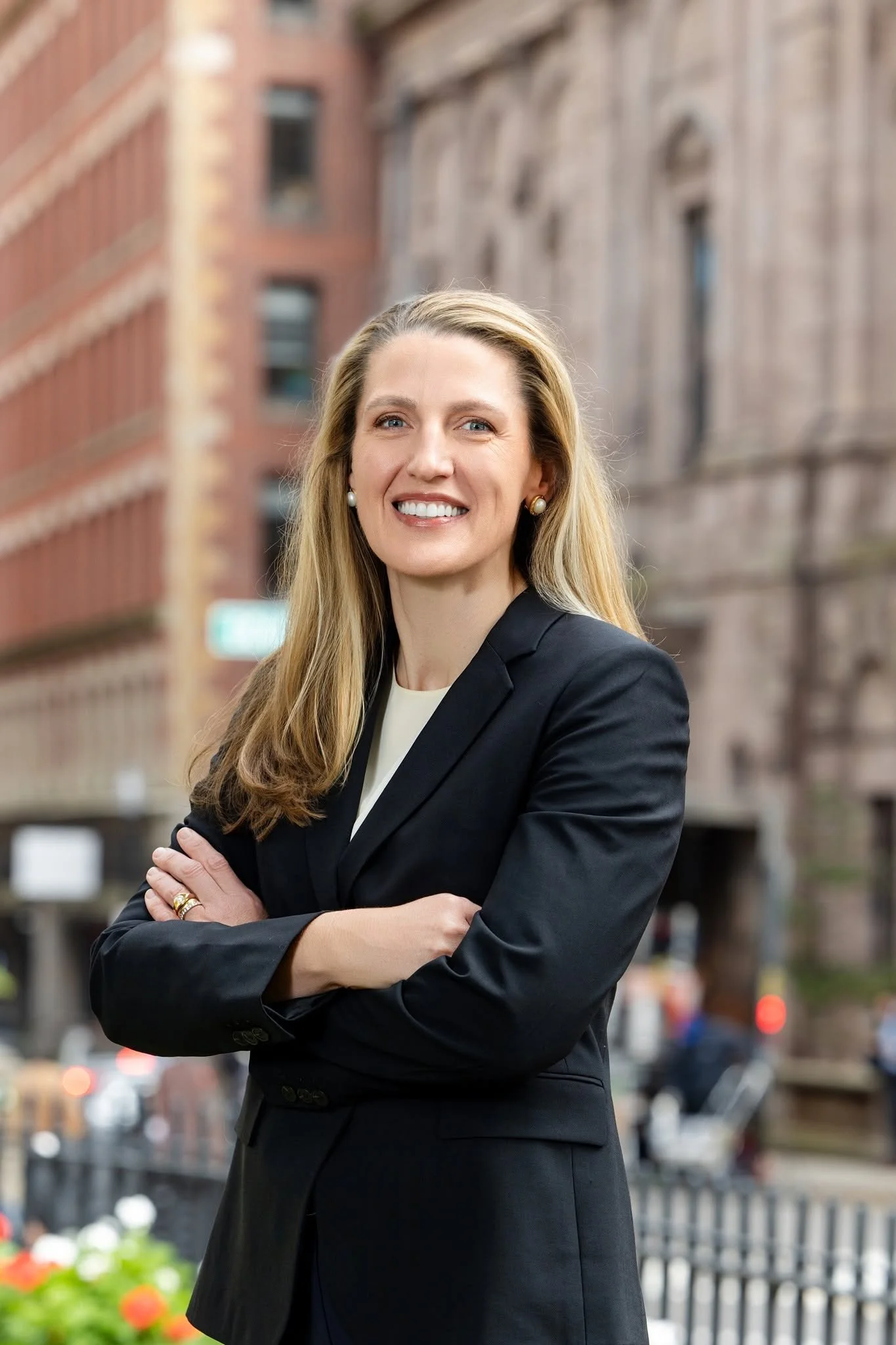 Portrait of Elizabeth Dionne, a smiling blonde woman with arms crossed, wearing a black blazer over a cream top, on a city street.