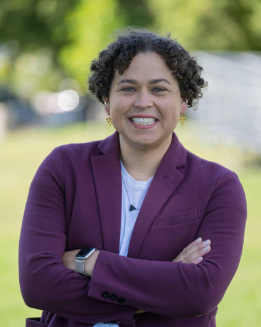Campaign photo of Carey McDonald, a person with short curly dark hair smiling outdoors with arms crossed, wearing a maroon blazer over a light top.