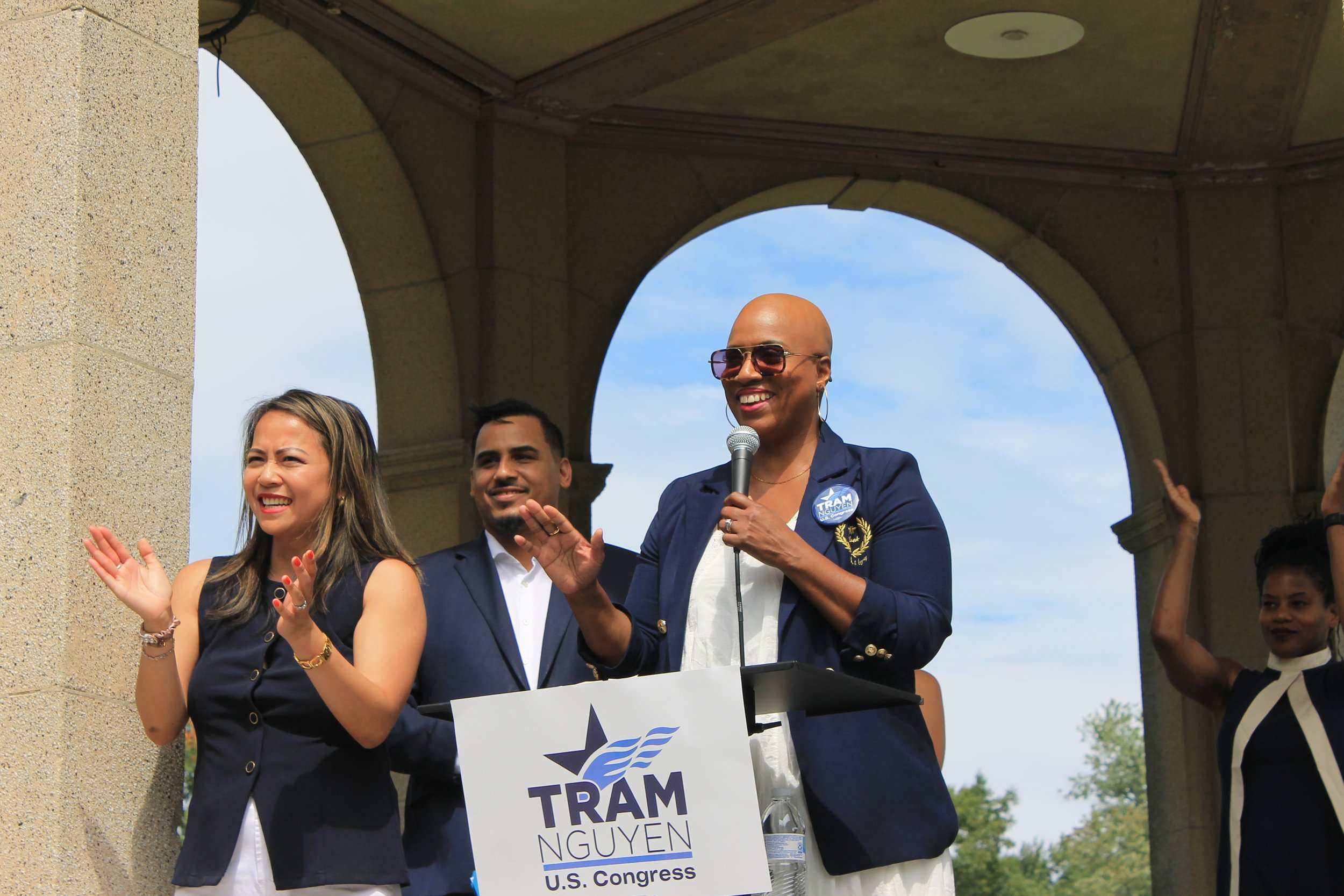 A speaker in a navy blazer and sunglasses speaks into a microphone beside Tram Nguyen, with supporters applauding around them.