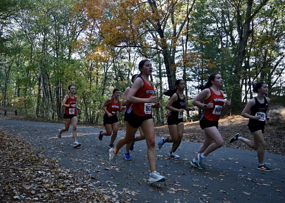 A pack of Reading girls cross-country runners in red uniforms competes along a leaf-covered path lined with autumn trees.