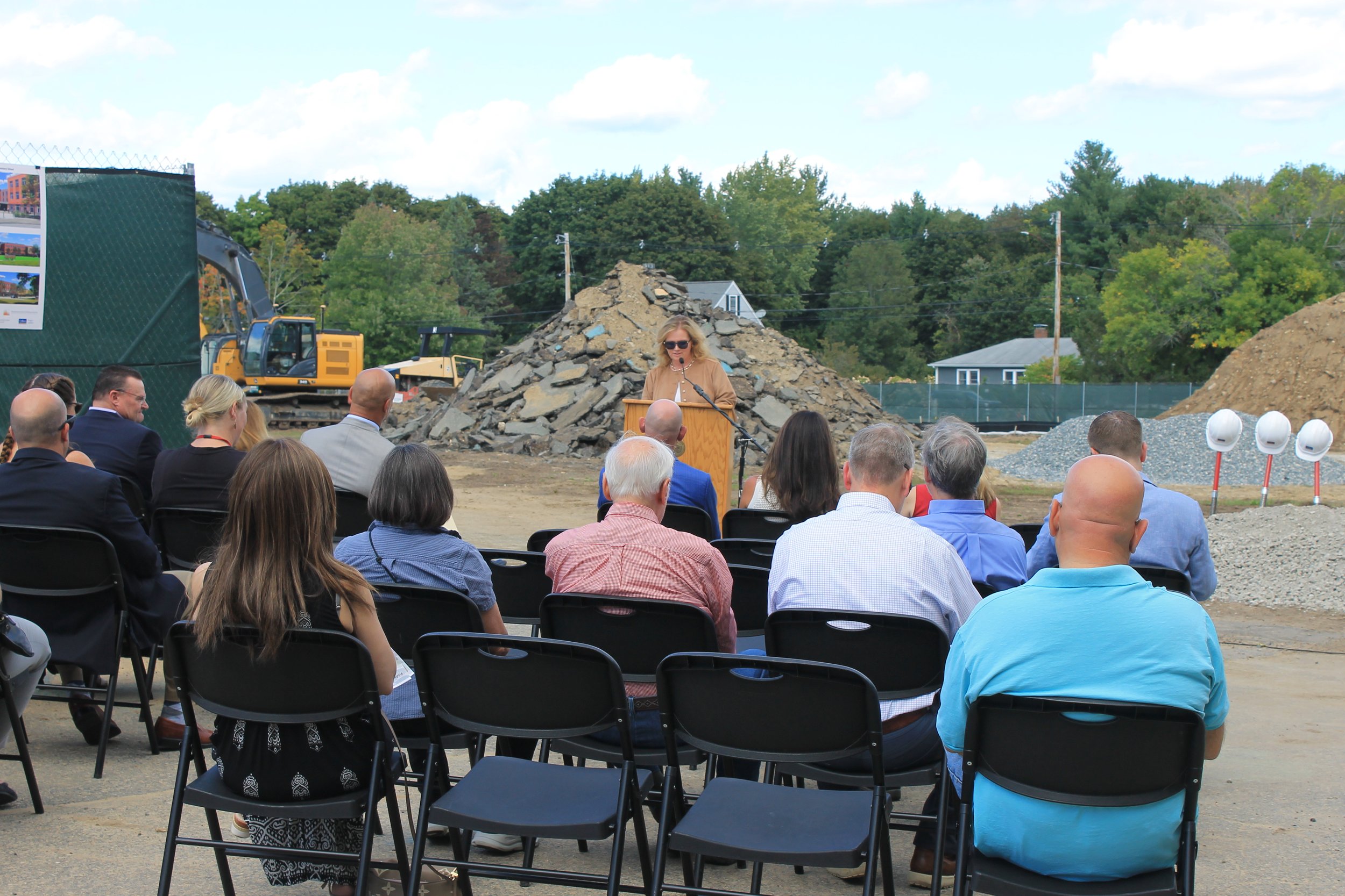 A speaker in a tan jacket addresses a seated crowd at a podium outdoors, with construction equipment, a debris pile, and ceremonial shovels and hard hats nearby.