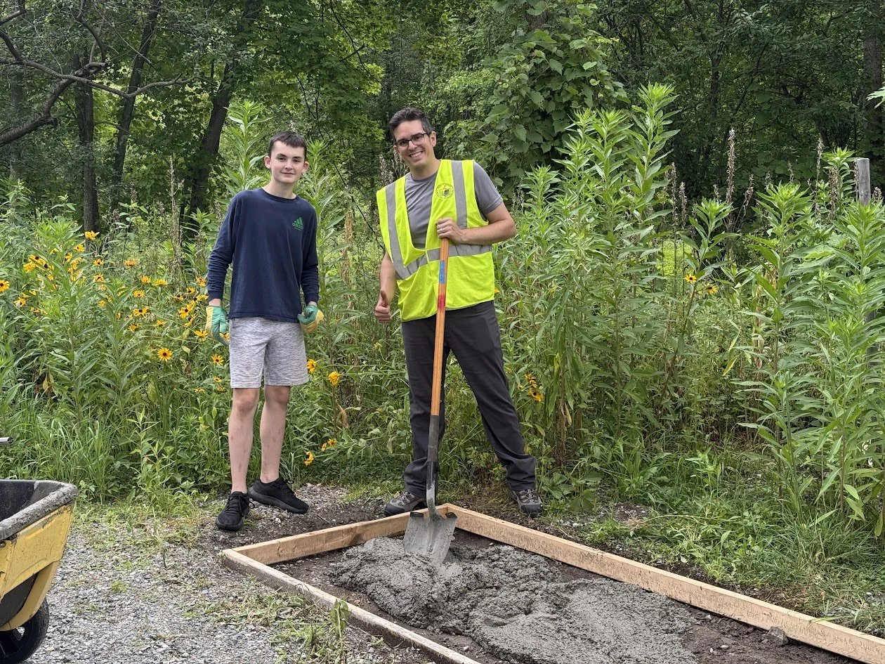 A young volunteer and an adult in a yellow safety vest stand behind a wooden form partly filled with concrete, a shovel resting in the mix.