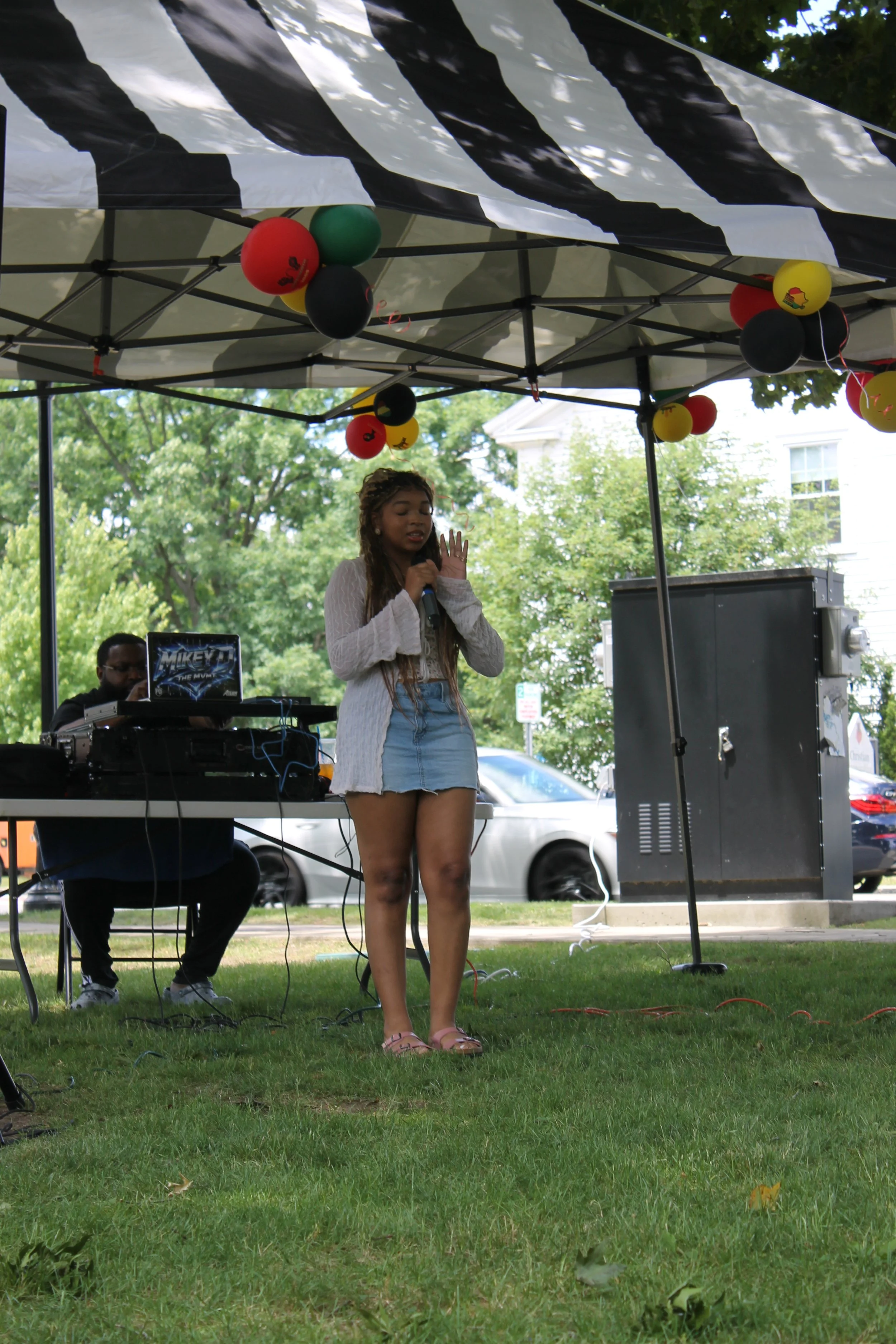 A girl sings into a microphone under a black-and-white striped tent, with red, green, black, and yellow balloons overhead and a DJ set up behind her.