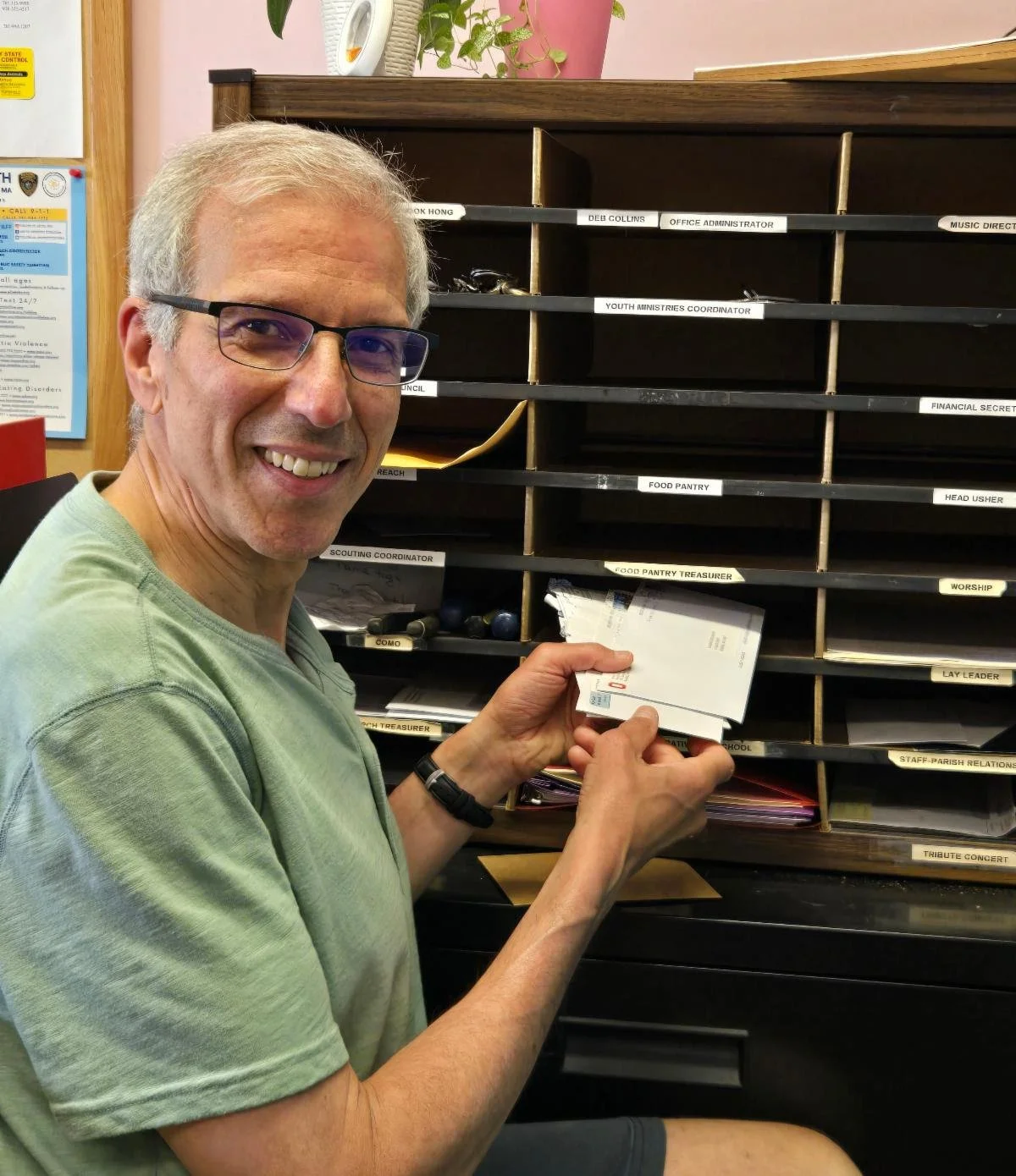 Hal Torman, wearing glasses and a green T-shirt, smiles while sorting mail at a wall-mounted mail sorter with labeled slots, including one for "Food Pantry Treasurer."