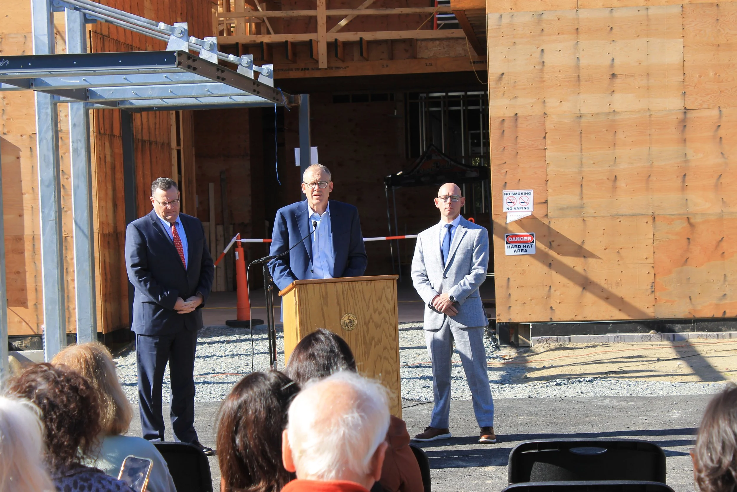 Man in a blue blazer speaks at a podium as two men in suits stand beside him before the wood-framed ReCAL building, with attendees in the foreground.