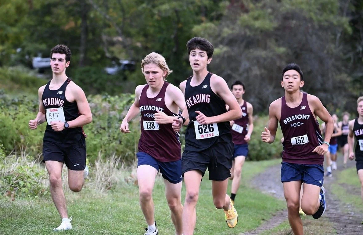 Two Reading boys' cross-country runners in black jerseys run alongside Belmont runners in maroon on a grassy course during a Middlesex League meet.