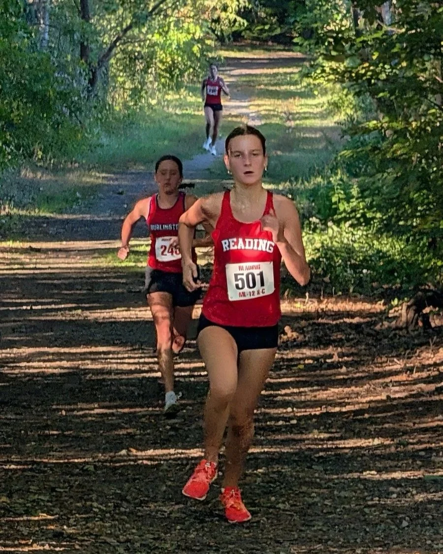 A Reading runner leads a Burlington runner on a wooded trail during a cross-country race, with another runner following behind.