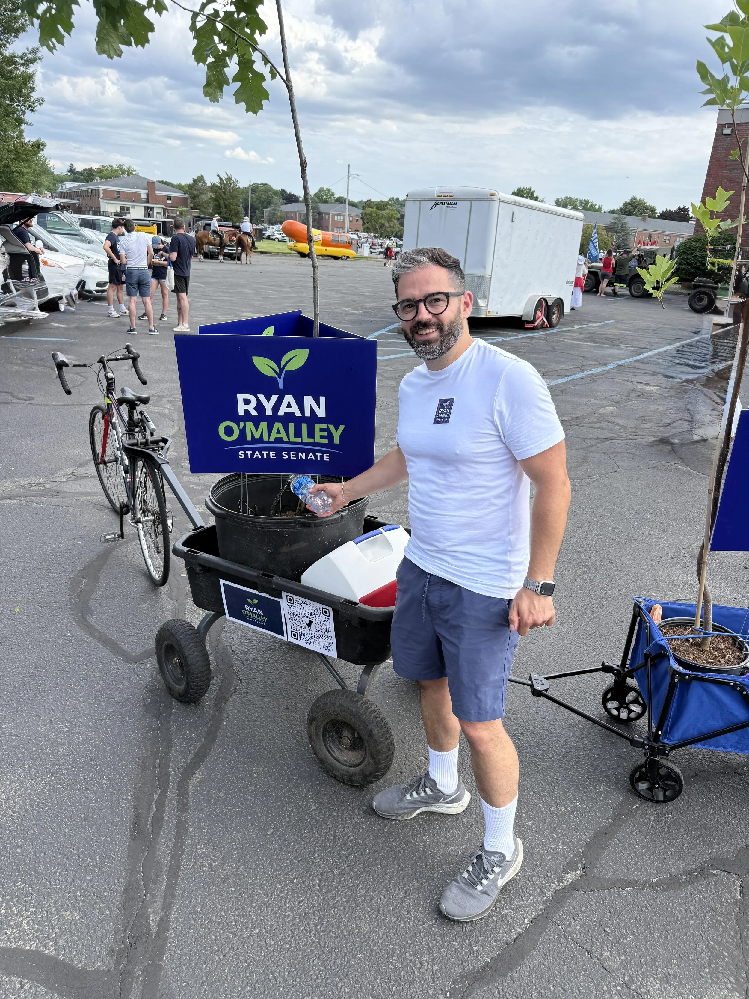 A man in glasses and a white t-shirt stands next to a wagon holding a "Ryan O'Malley State Senate" sign and a young potted tree, at an outdoor community event with vehicles and a hot dog-shaped car in the background.