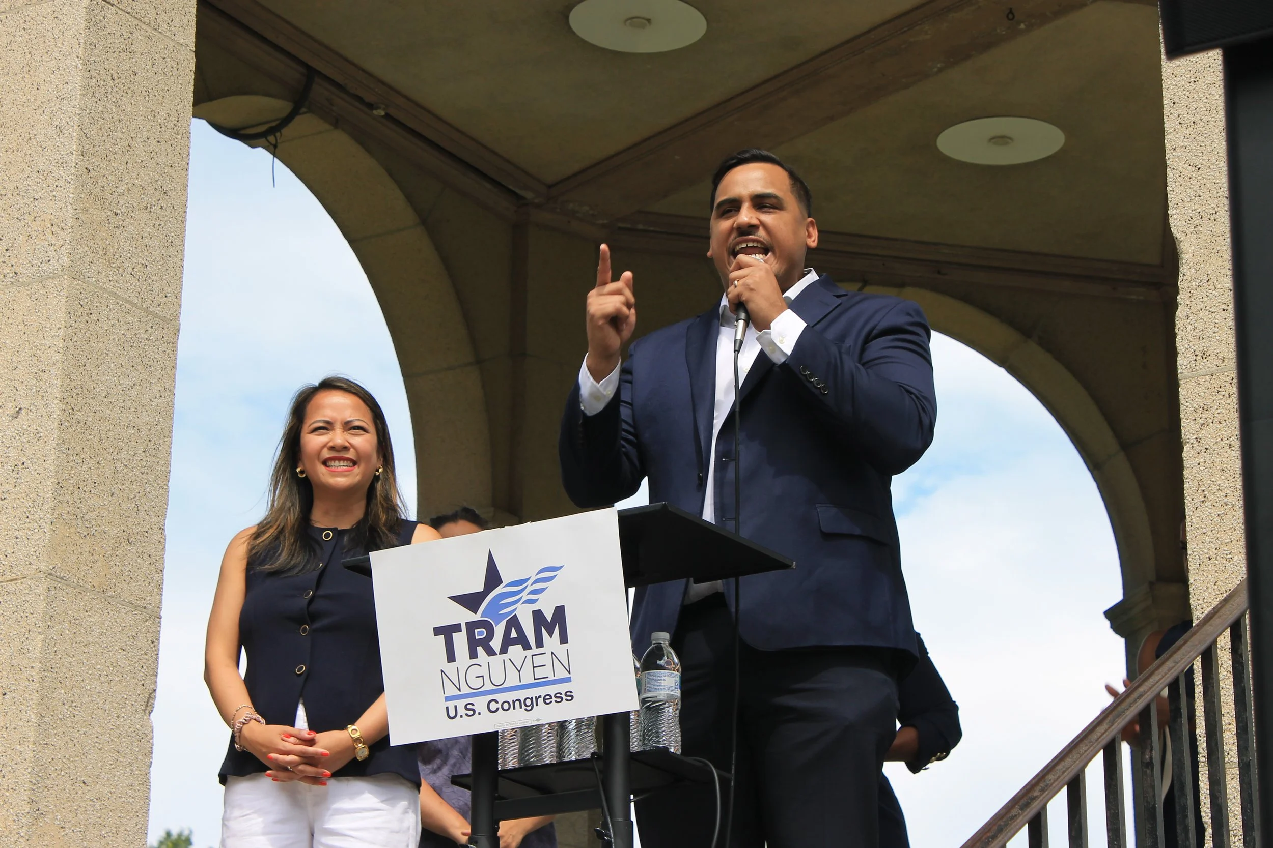 A man in a navy suit speaks at a podium with a "Tram Nguyen for U.S. Congress" sign, gesturing with one hand raised, as Nguyen stands beside him smiling.